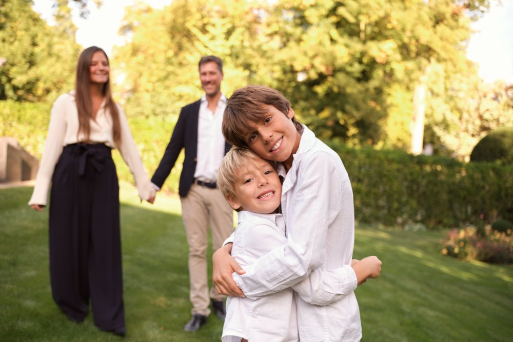 Two brothers hugging in a garden while their parents walk behind them